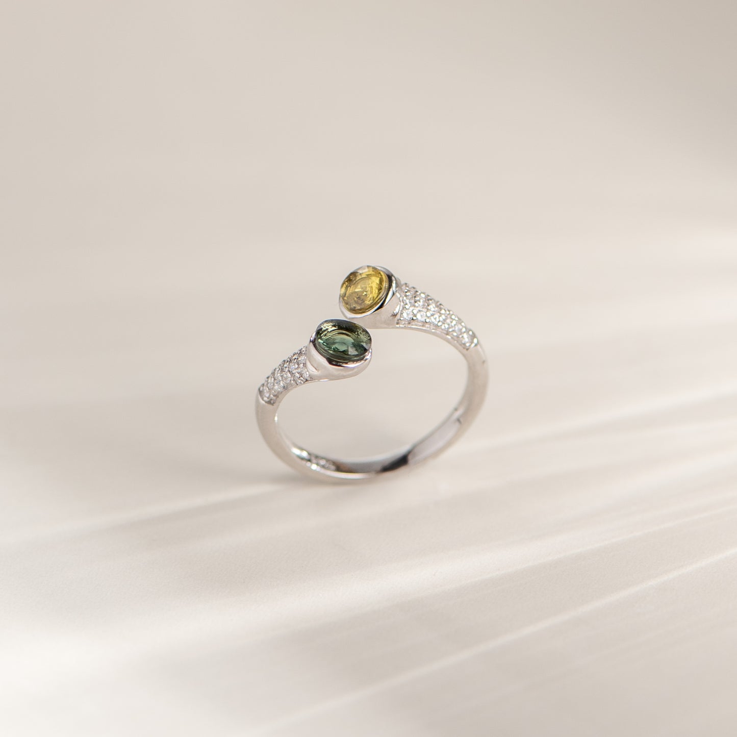 A silver ring with two oval gemstones, one yellow and one green, accented by small clear stones on a light beige background.