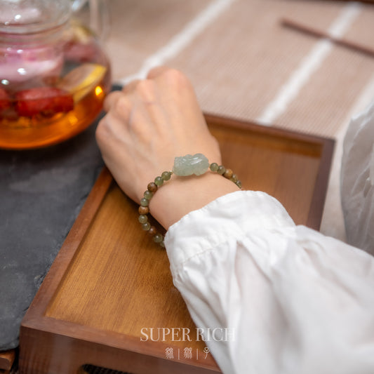 Hand wearing a bracelet with a stone charm on a wooden surface next to a teapot.