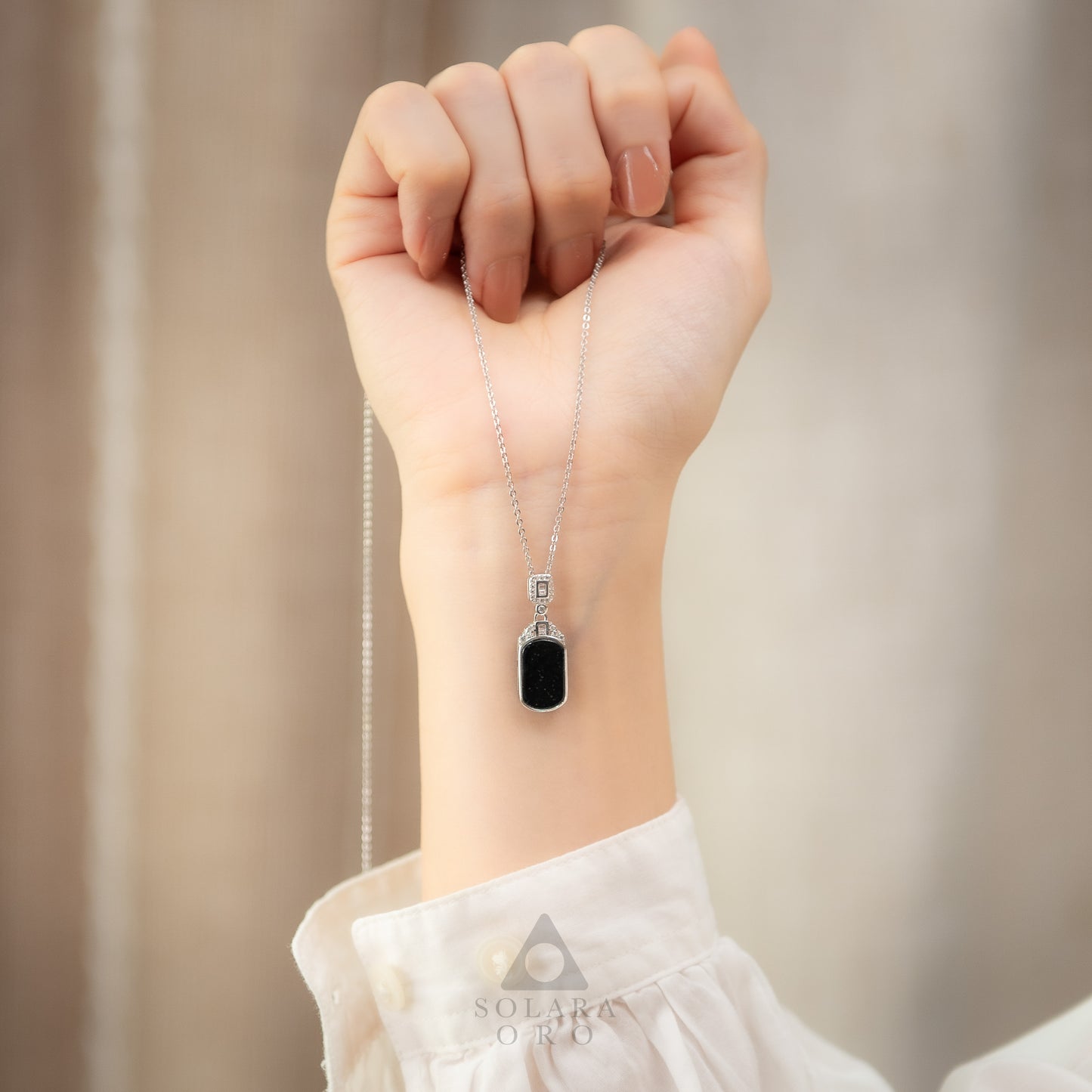 Hand wearing a necklace with a black pendant against a neutral background