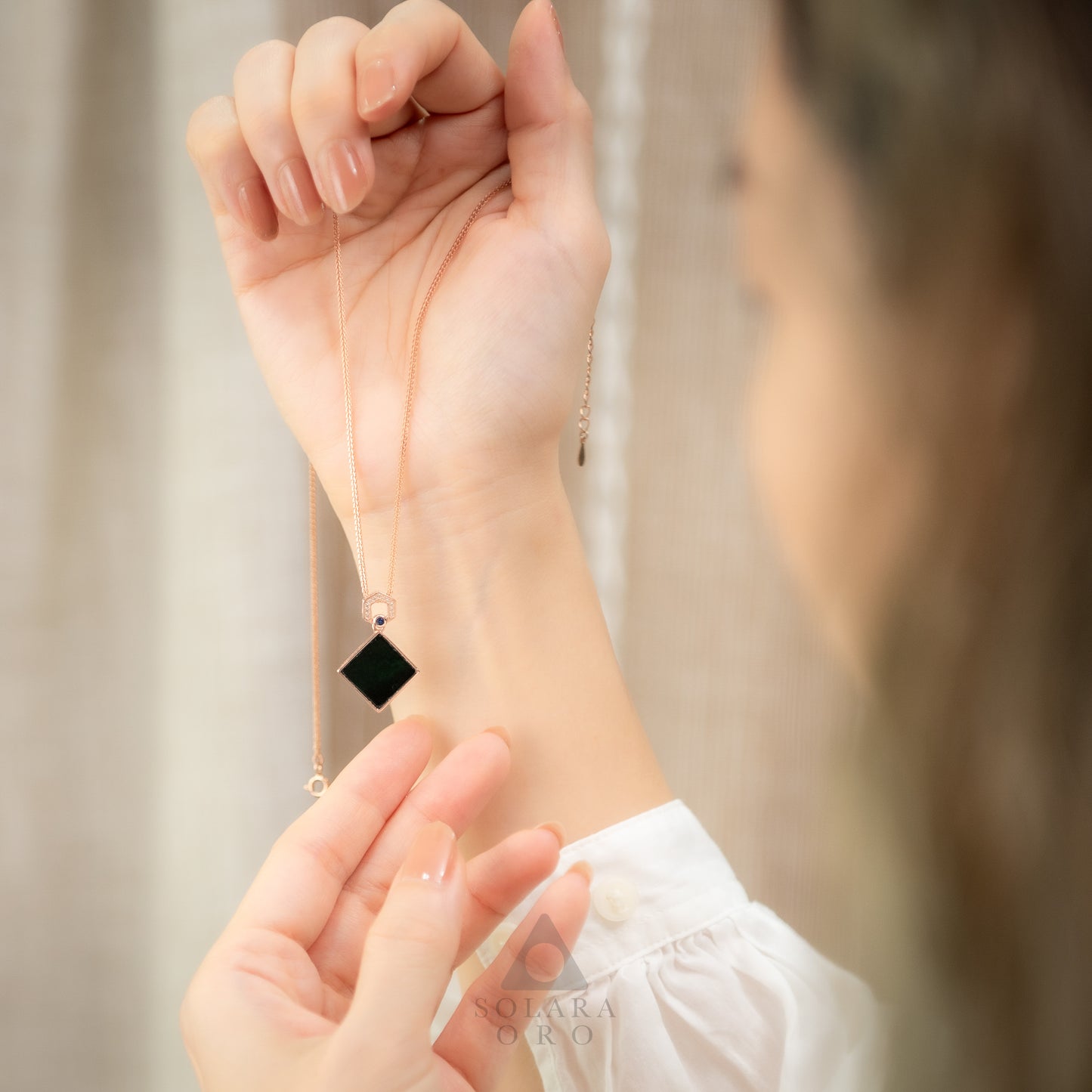 Person wearing a black square pendant on a delicate chain, with a blurred background.