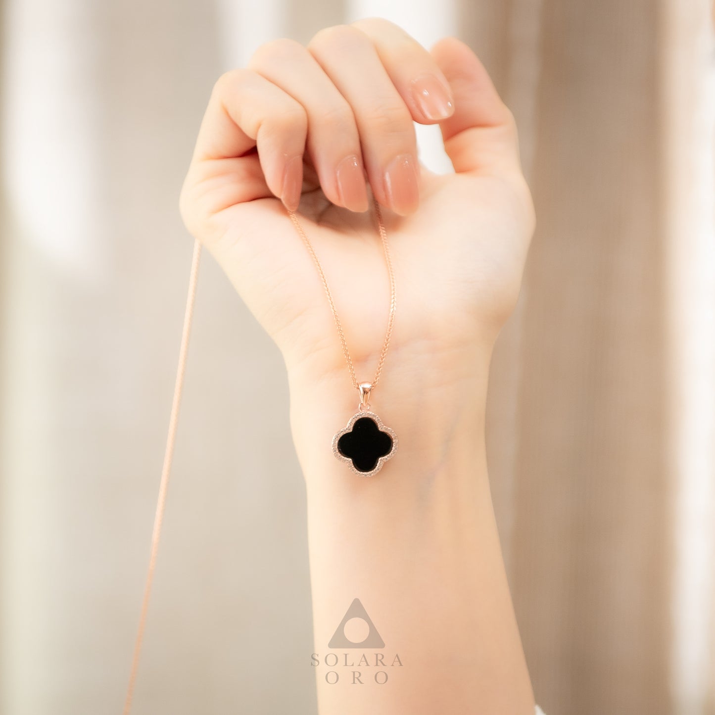 Hand holding a necklace with a black clover pendant against a neutral background