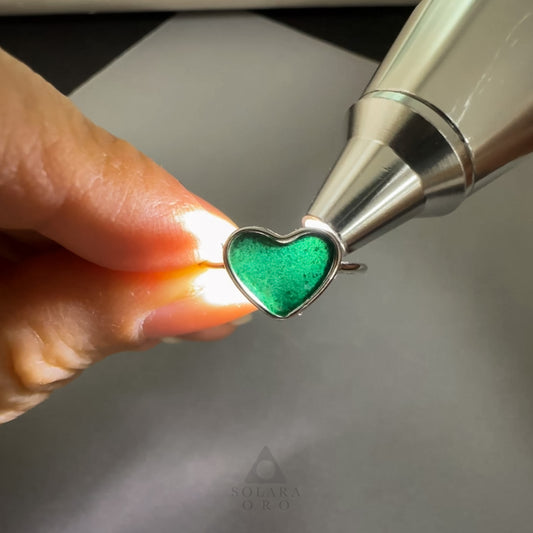 Heart-shaped green gemstone being inserted into a silver setting by a hand.