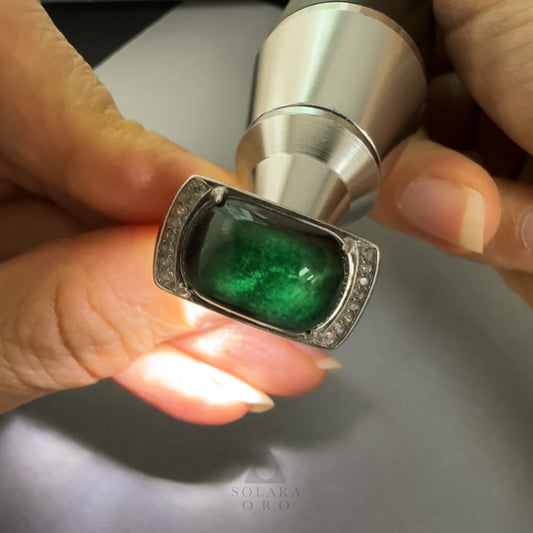 Close-up of a hand holding a silver ring with a green gemstone on a neutral background.
