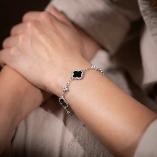 A close-up of a woman wearing a Solara Oro silver clover bracelet with black stone accents, resting her hands together in a calm, intimate moment.