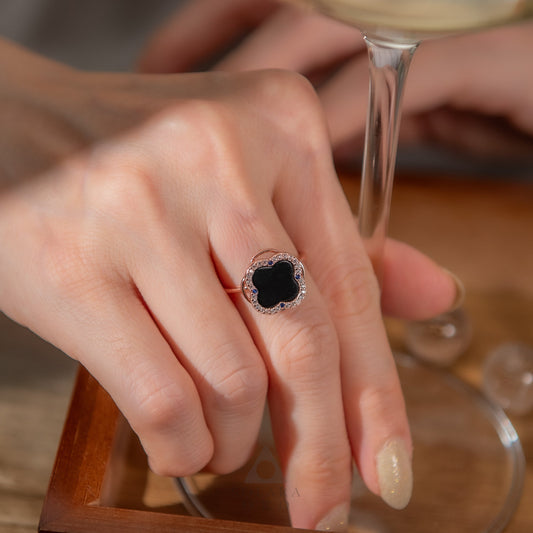 Close-up of a woman’s hand wearing a black clover gemstone ring with diamond halo, resting beside a wine glass in warm natural light, symbolizing elegance and grounding energy.