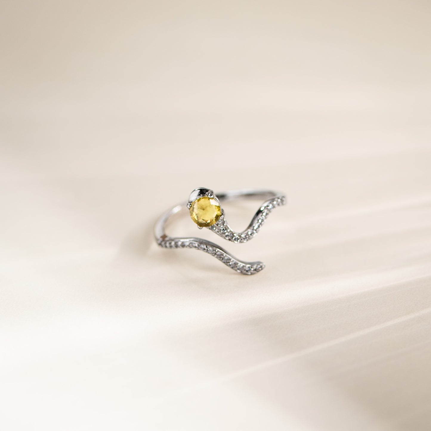 A silver wave-shaped ring with a central oval yellow gemstone and small clear stones along the band, photographed on a soft cream background.