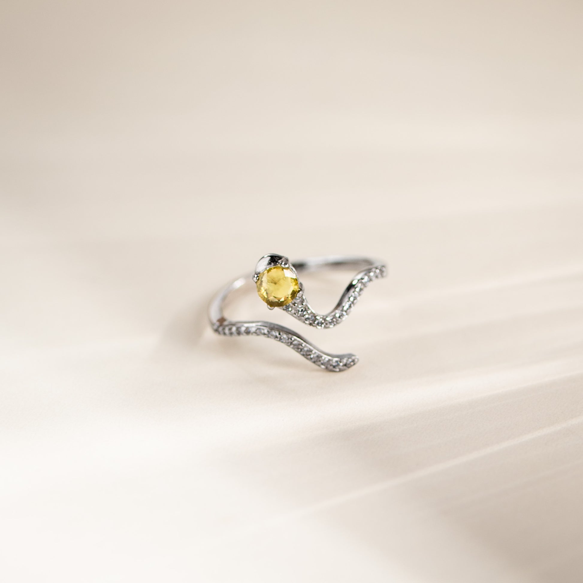 A silver wave-shaped ring with a central oval yellow gemstone and small clear stones along the band, photographed on a soft cream background.