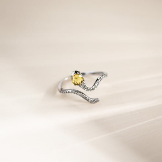 A silver wave-shaped ring with a central oval yellow gemstone and small clear stones along the band, photographed on a soft cream background.