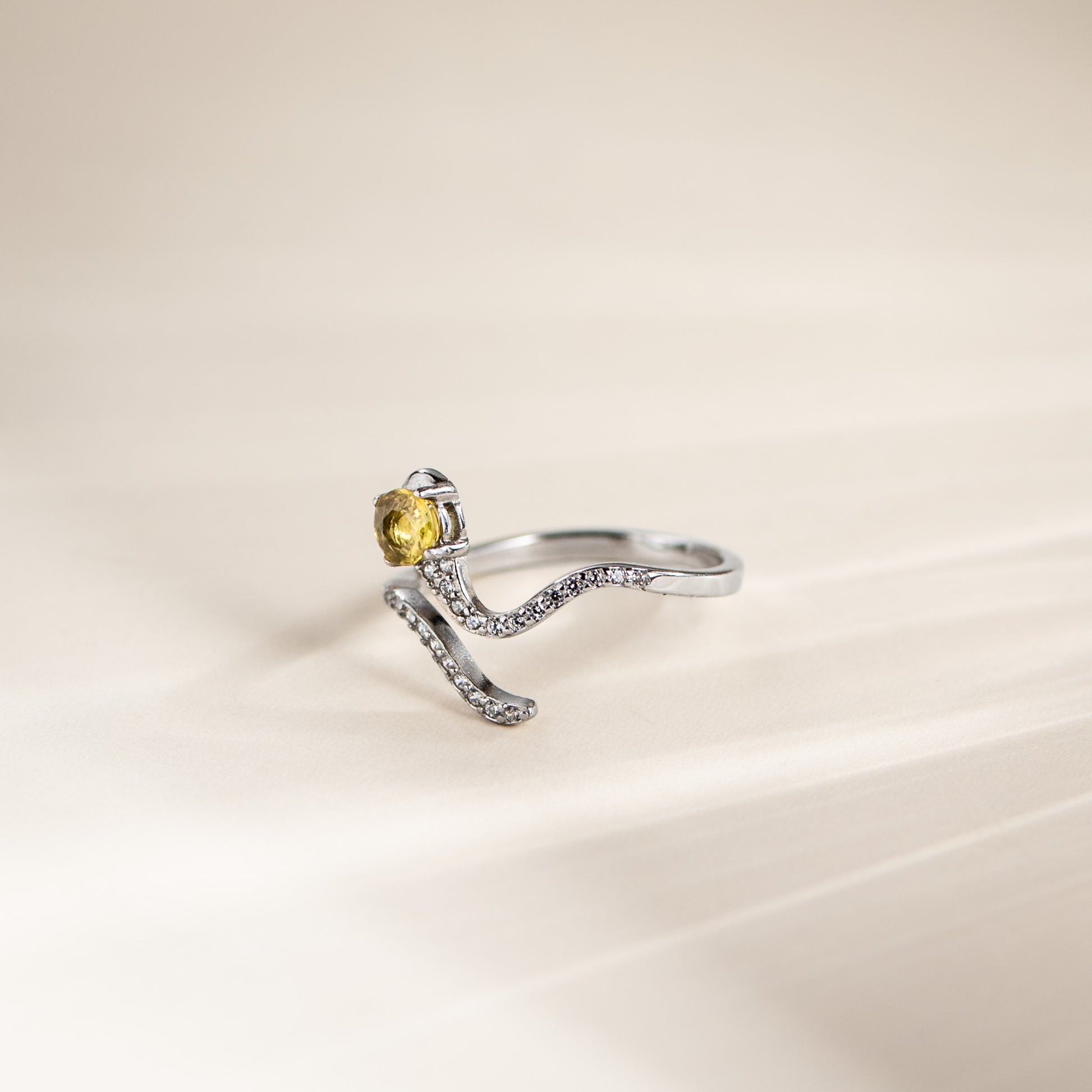 A silver-toned open ring with a curved, pavé-set band and a round yellow gemstone, photographed on a soft cream background.