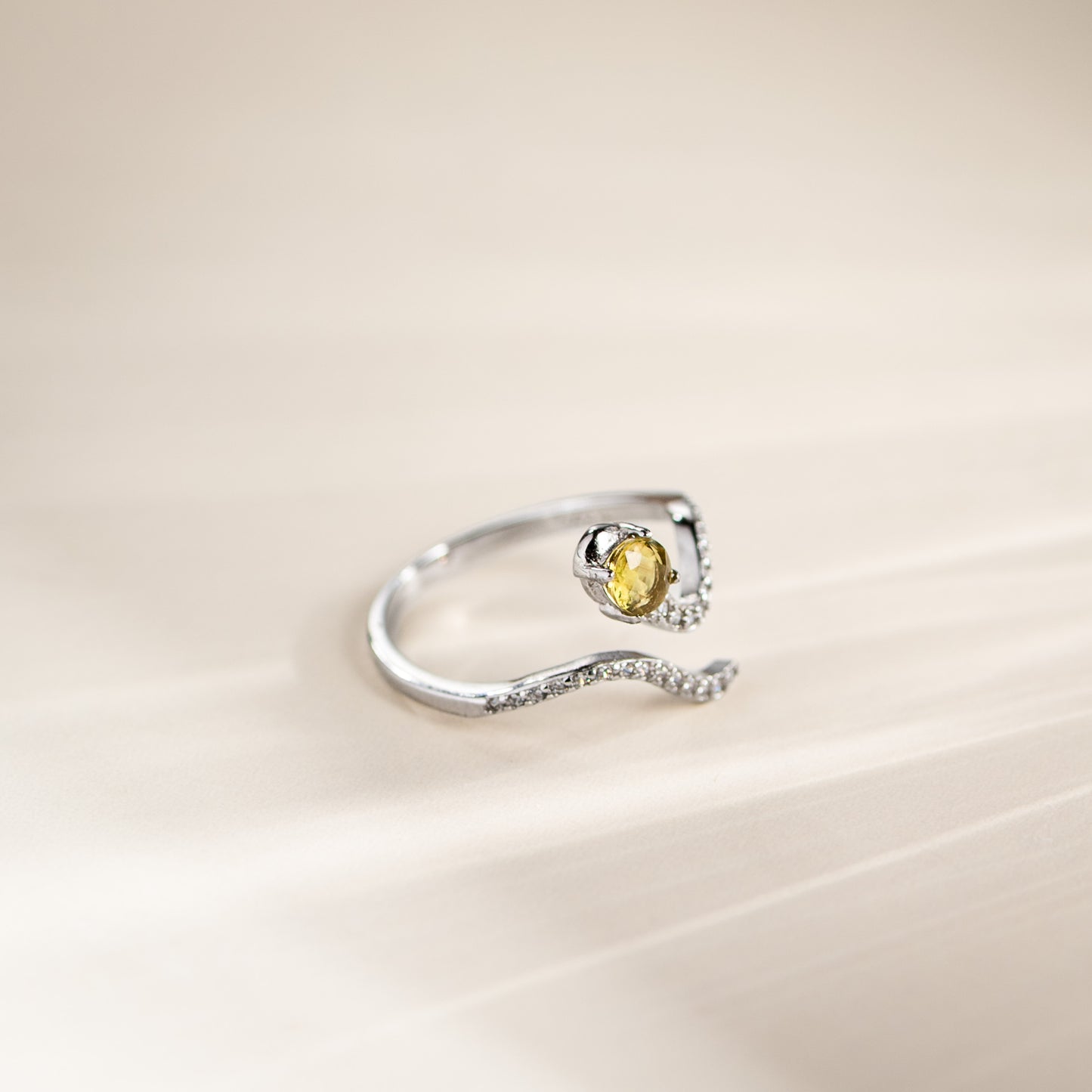 A silver open-band ring featuring a curved, wavy band set with small clear stones and a central oval yellow gemstone, photographed on a soft beige background.