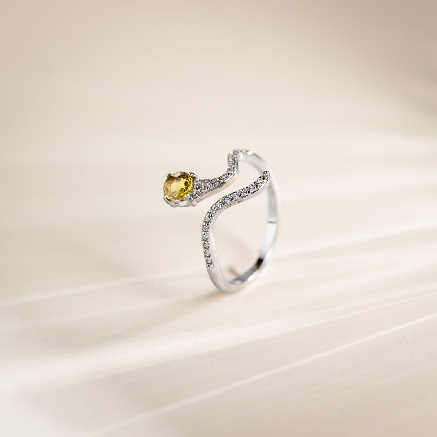 A silver ring featuring a curved, pavé-set band and a round-cut yellow gemstone, photographed on a soft beige background.