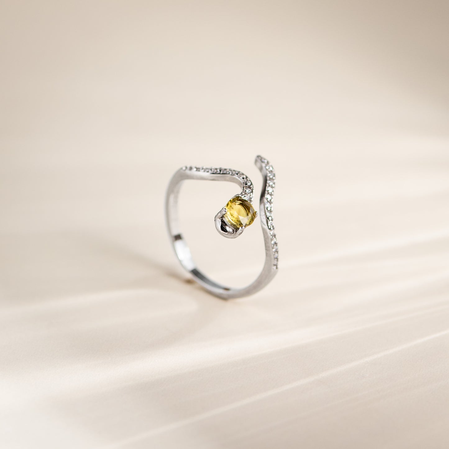 A silver-toned open ring with a curved, pavé-set band and a central round yellow gemstone, photographed on a soft beige background.