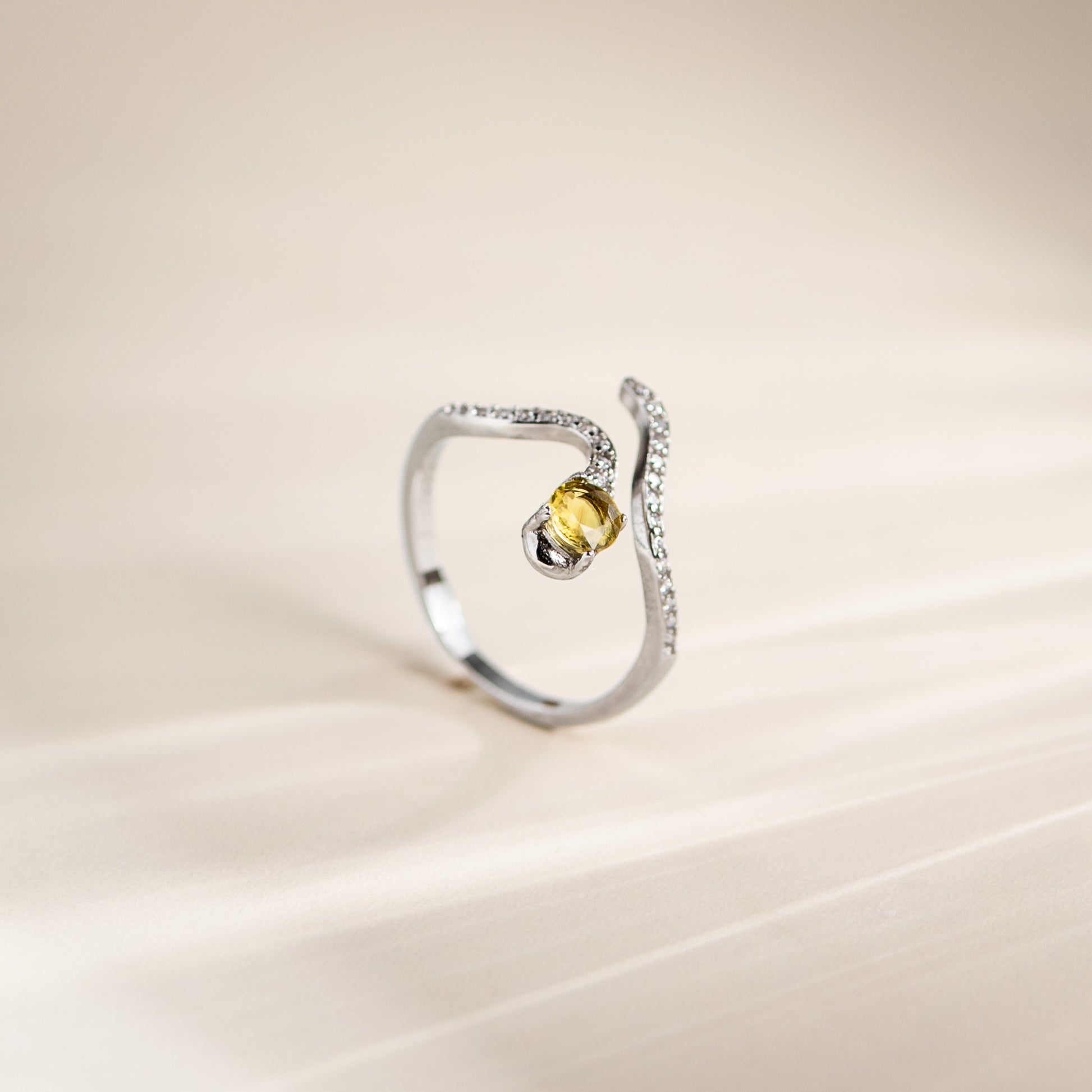 A silver-toned open ring with a curved, pavé-set band and a central round yellow gemstone, photographed on a soft beige background.