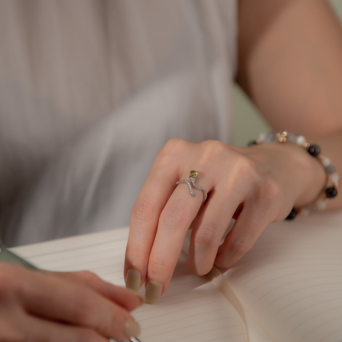 A close-up lifestyle shot of a person's hand resting on an open notebook, wearing a silver wavy ring with a small green gemstone and a beaded bracelet.