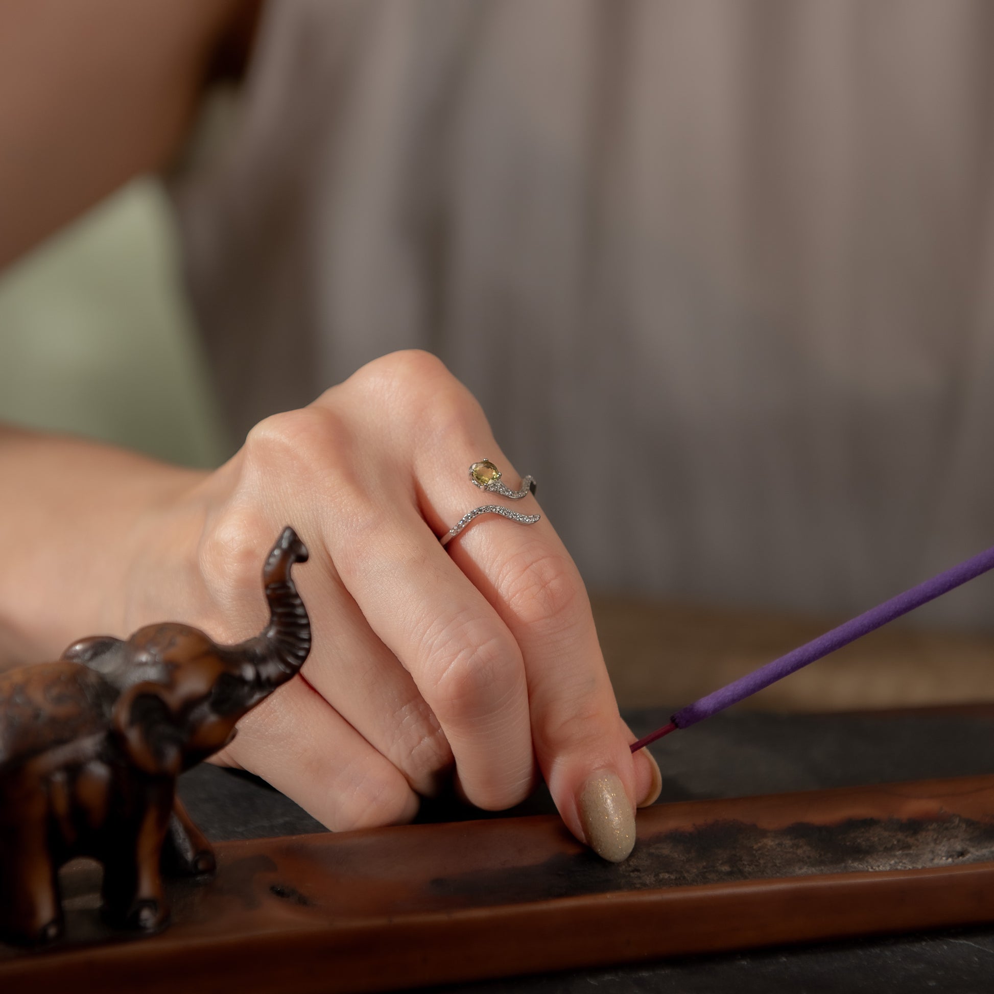 A close-up lifestyle shot of a person's hand wearing a silver snake ring with a small yellow gemstone, resting on a wooden desk next to a small elephant figurine.