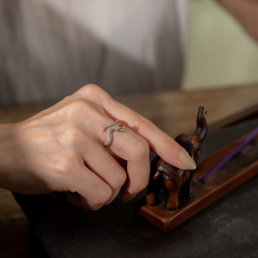 A close-up of a hand wearing a snake-shaped ring with a small yellow gemstone, resting near a small elephant figurine on a wooden stand.