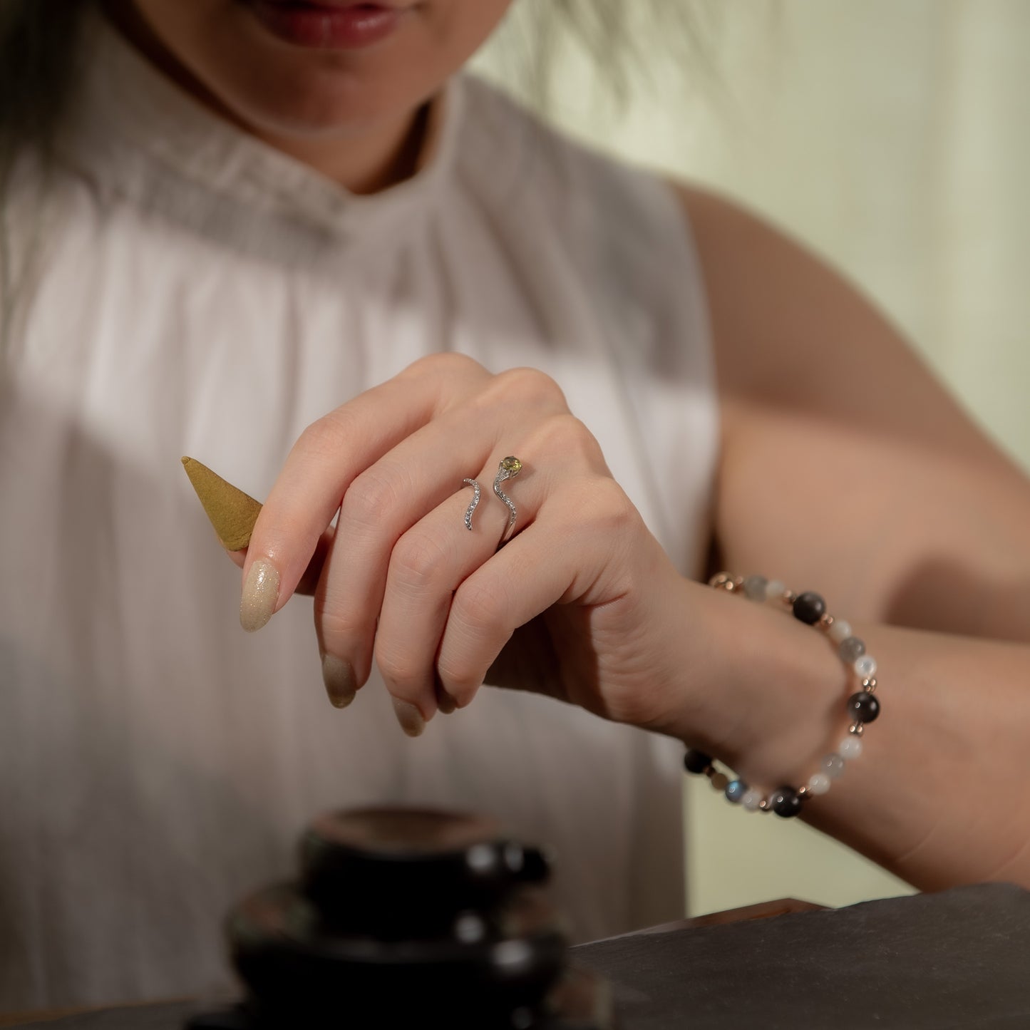 A close-up lifestyle shot of a woman's hand wearing a silver snake-shaped ring with a green stone and a beaded bracelet, while holding a small gold object.