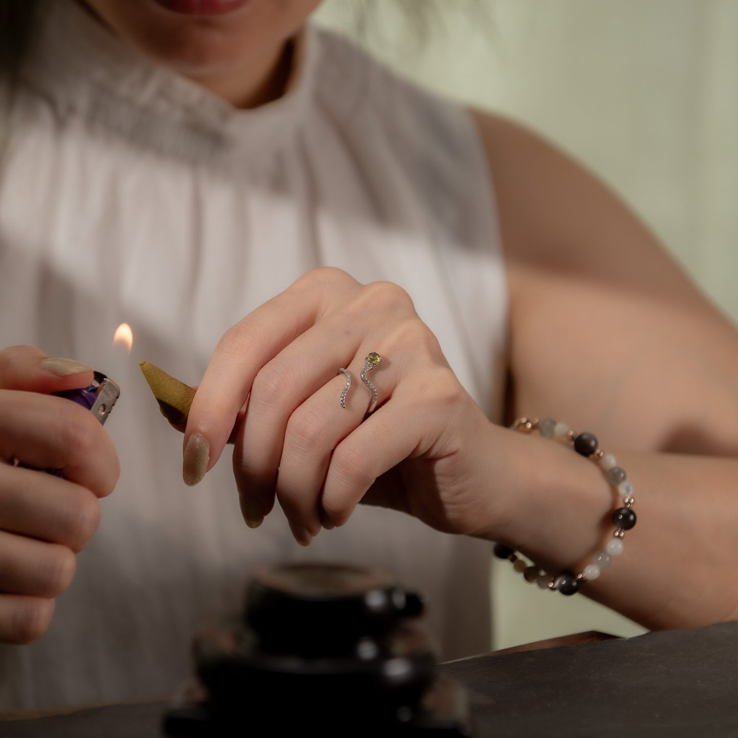 A close-up lifestyle shot of a woman's hands lighting a incense stick, wearing a silver snake-shaped ring and a beaded bracelet.