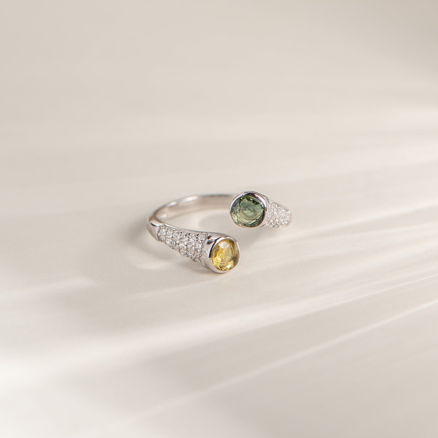 A silver open ring ring featuring one oval green gemstone and one oval yellow gemstone, both accented with small pavé-set clear stones, resting on a light beige surface.