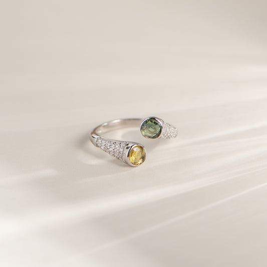 A silver open ring ring featuring one oval green gemstone and one oval yellow gemstone, both accented with small pavé-set clear stones, resting on a light beige surface.