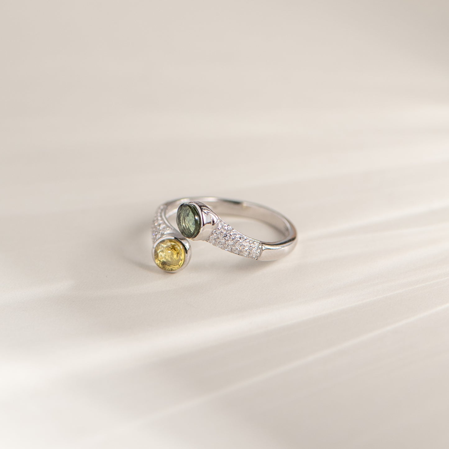 A silver wrap-style ring featuring one oval green gemstone and one round yellow gemstone, accented with small clear stones along the band, photographed on a soft cream background.