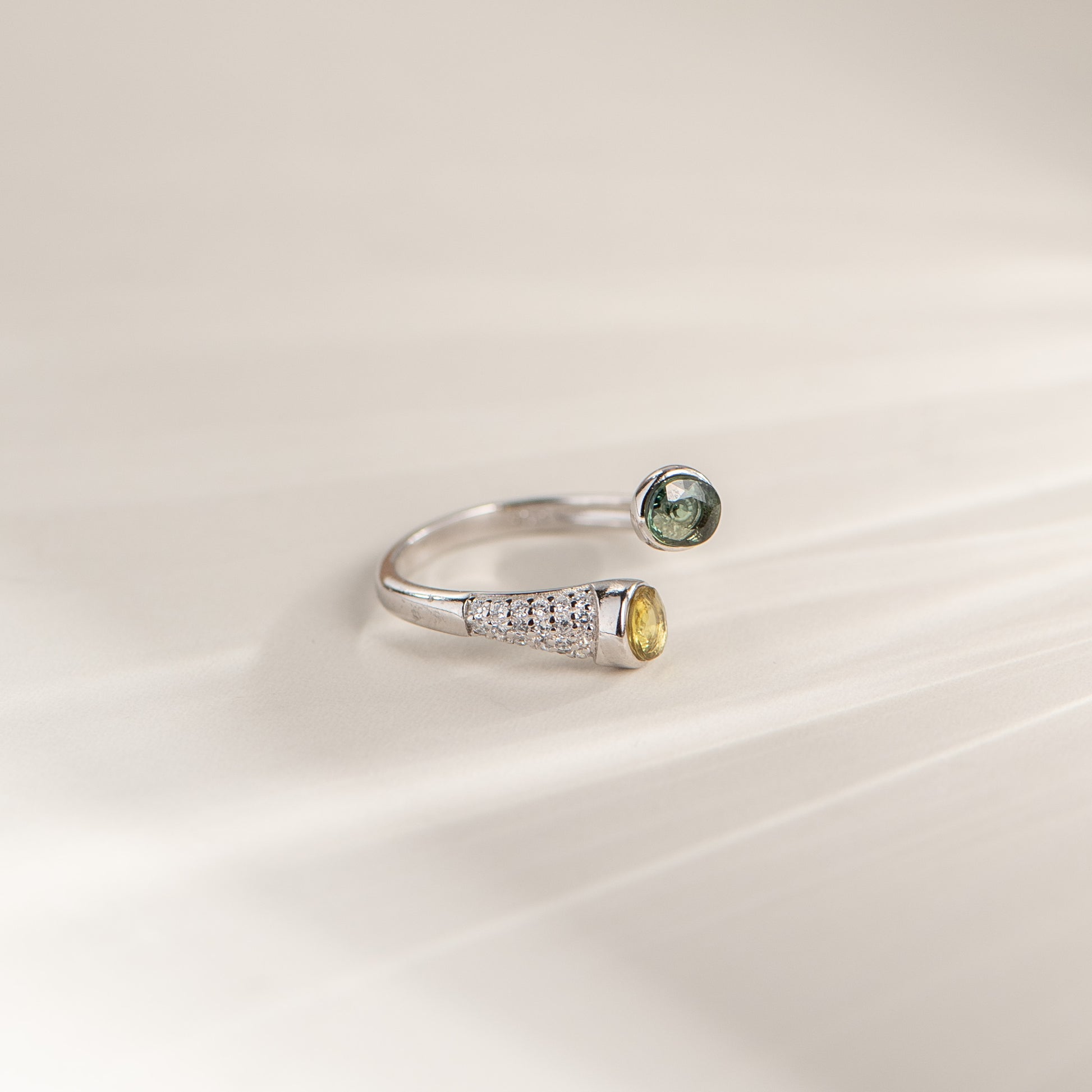 A silver open ring band featuring a pavé-set clear stone section and a yellow gemstone on one end, paired with a round green gemstone on the other, photographed on a soft cream background.