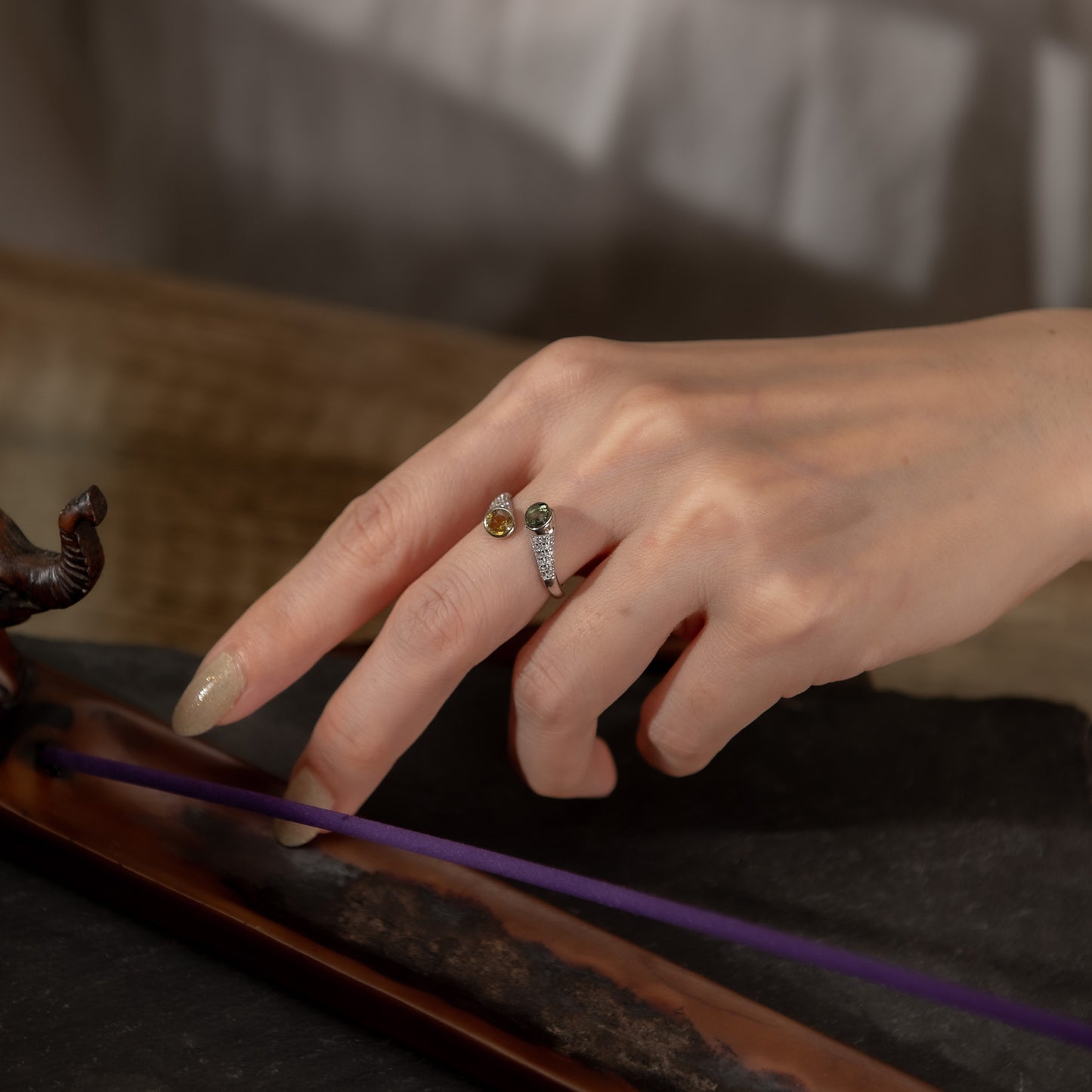 A close-up of a person's hand wearing a silver ring with small gold and green stones, resting near a purple incense stick on a dark surface.