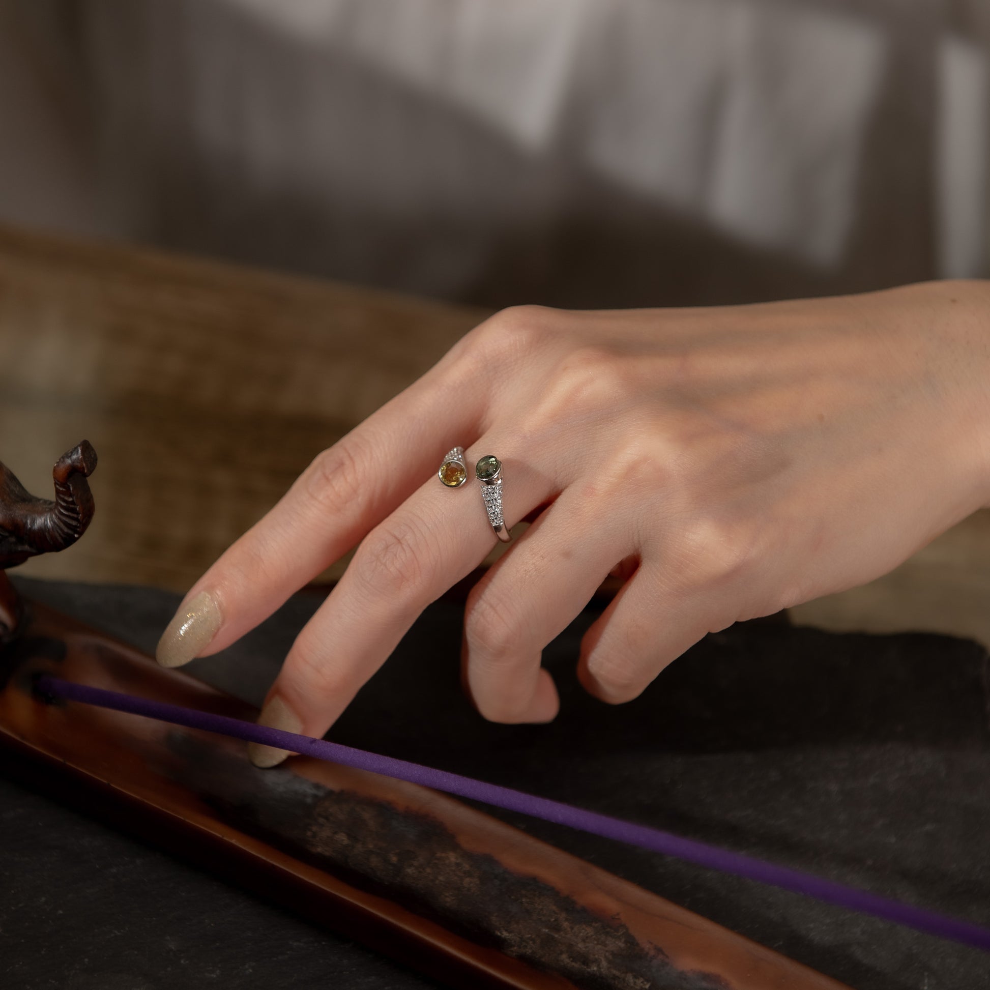 A close-up of a person's hand wearing a silver ring with small gold and green stones, resting near a purple incense stick on a dark surface.