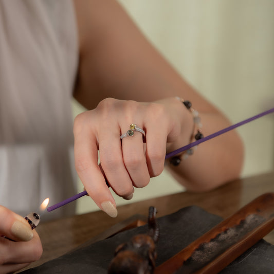 A close-up lifestyle shot of a person's hands lighting a purple incense stick with a lighter. The person is wearing a silver ring with a small green and yellow stone on the ring finger, and a beaded bracelet on the wrist.