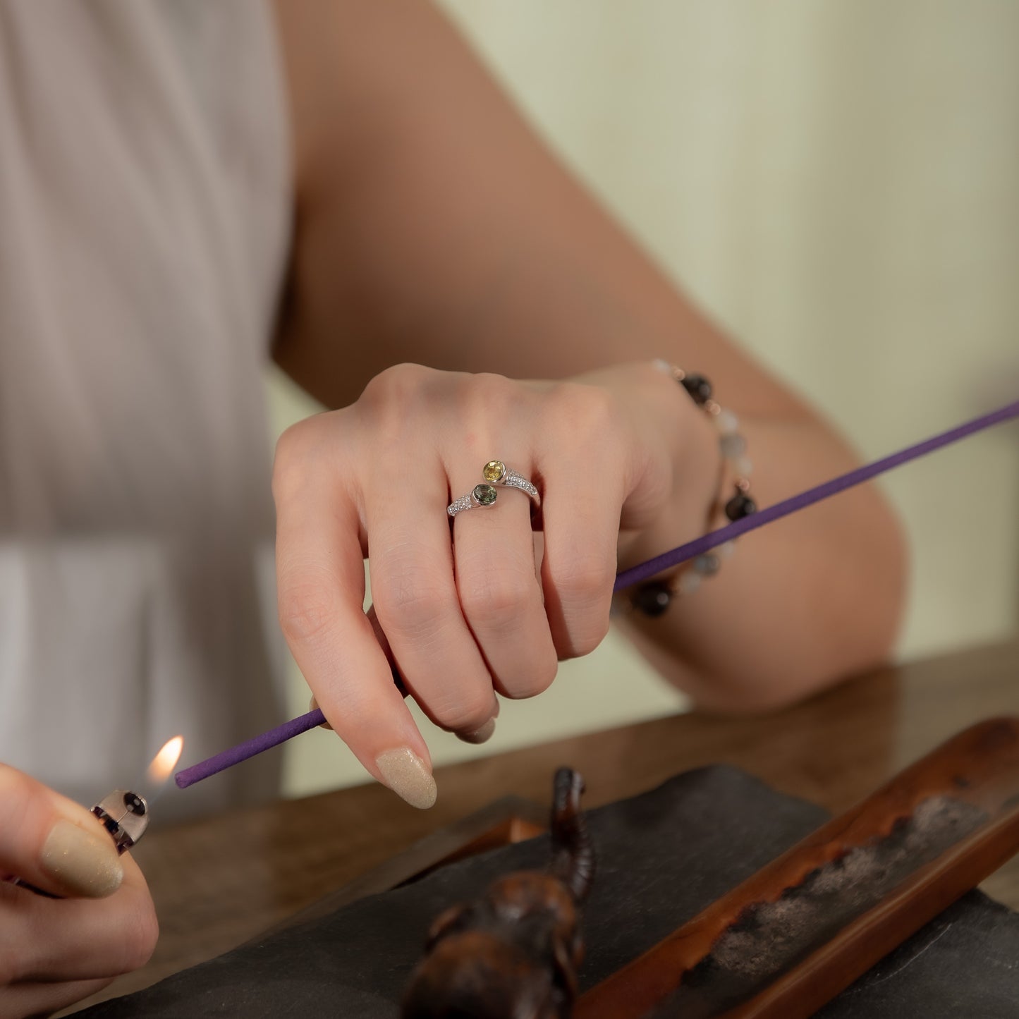A close-up lifestyle shot of a person's hands lighting a purple incense stick with a lighter. The person is wearing a silver ring with a small green and yellow stone on the ring finger, and a beaded bracelet on the wrist.