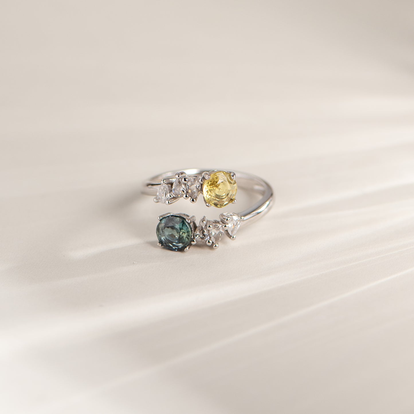 A silver-colored ring featuring a yellow gemstone, a blue gemstone, and several small clear accent stones, displayed on a soft, light beige background.