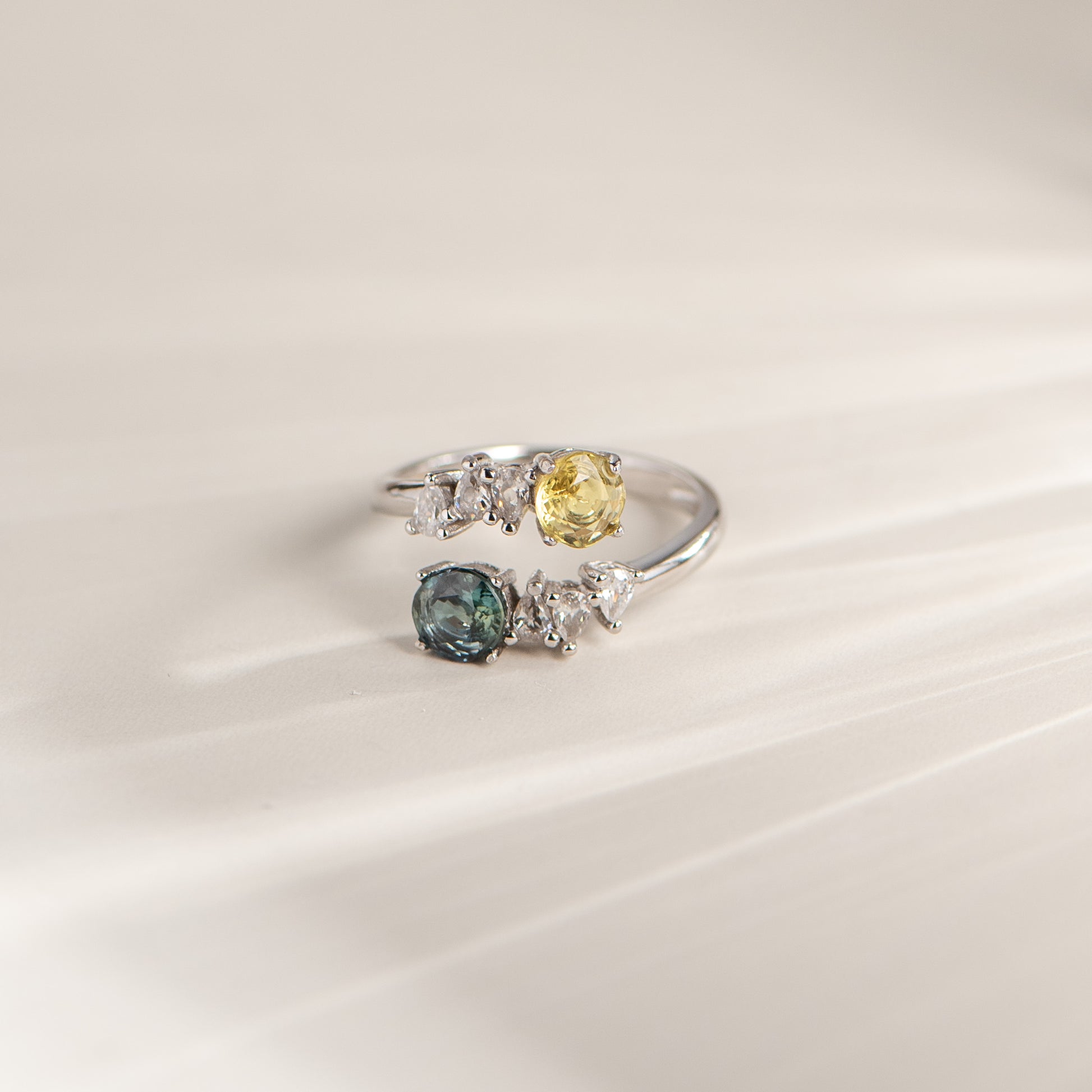 A silver-colored ring featuring a yellow gemstone, a blue gemstone, and several small clear accent stones, displayed on a soft, light beige background.