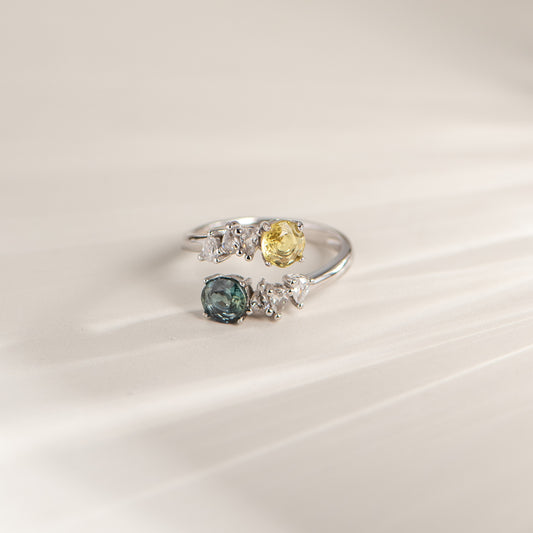 A silver-colored ring featuring a yellow gemstone, a blue gemstone, and several small clear accent stones, displayed on a soft, light beige background.