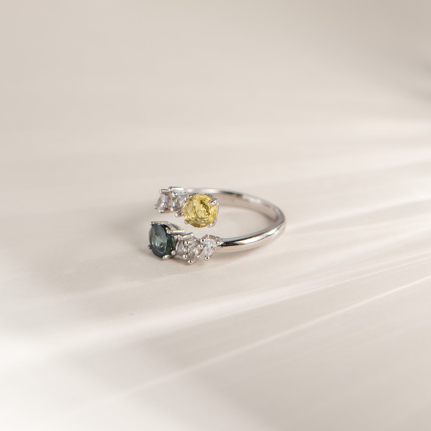 A silver ring featuring a yellow round-cut gemstone and a smaller dark blue-green oval-cut gemstone, each flanked by small clear stones, set against a soft beige background.