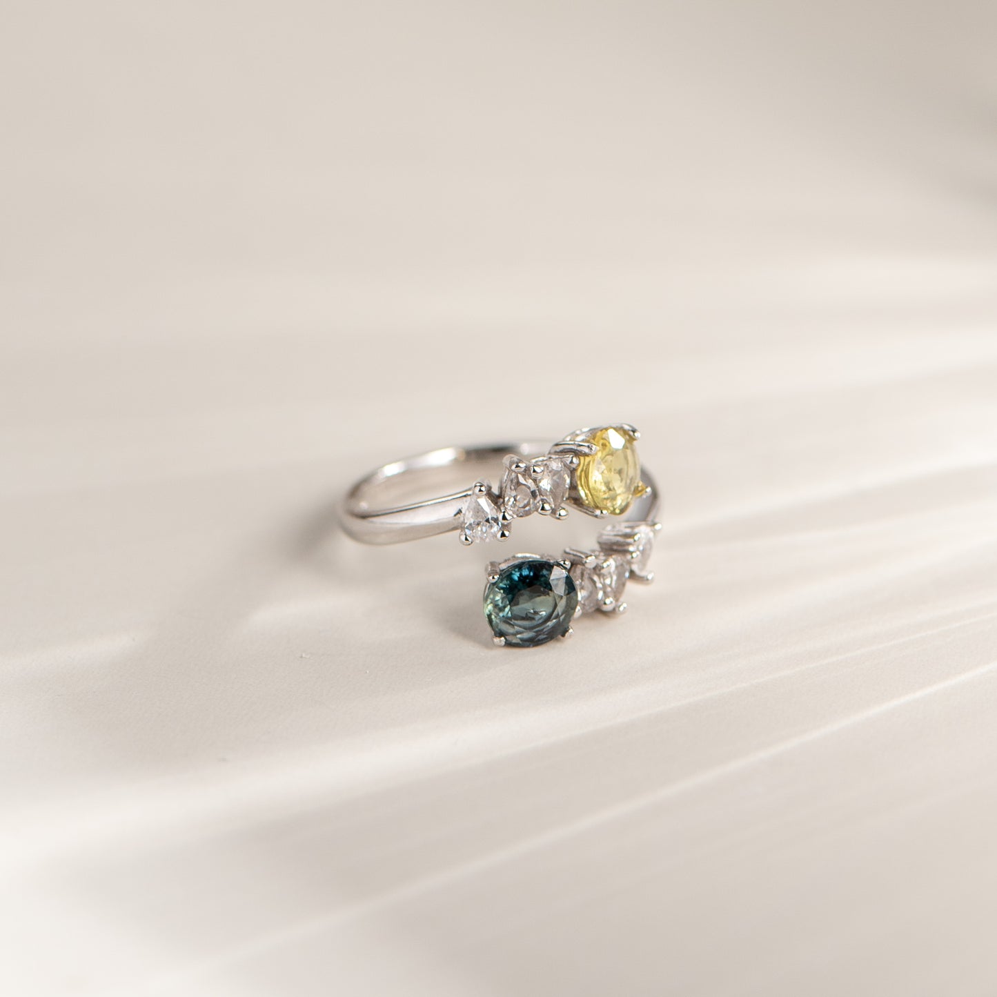 A silver ring with a yellow gemstone and a smaller blue gemstone, photographed on a soft, light beige background with a subtle diagonal shadow.