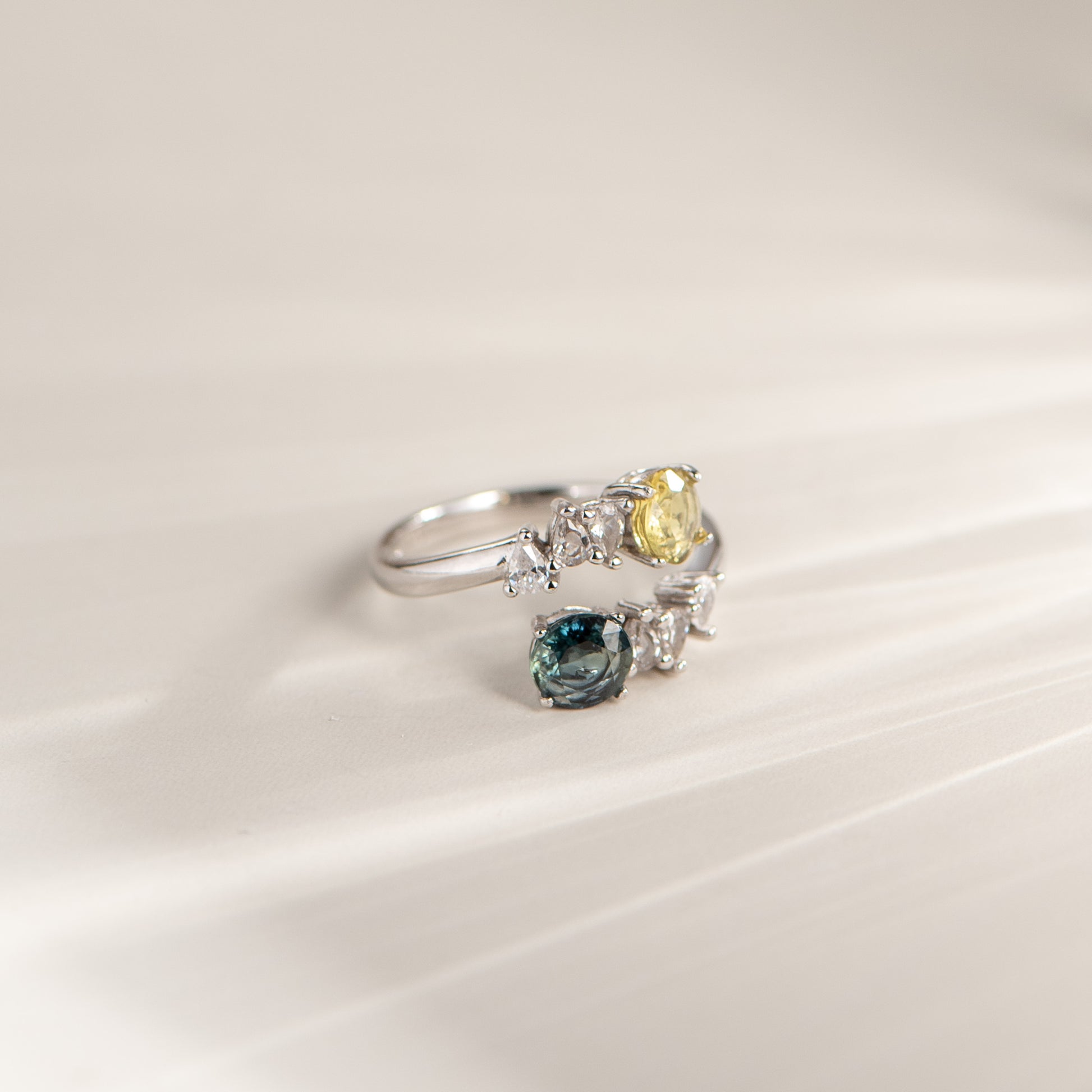 A silver ring with a yellow gemstone and a smaller blue gemstone, photographed on a soft, light beige background with a subtle diagonal shadow.