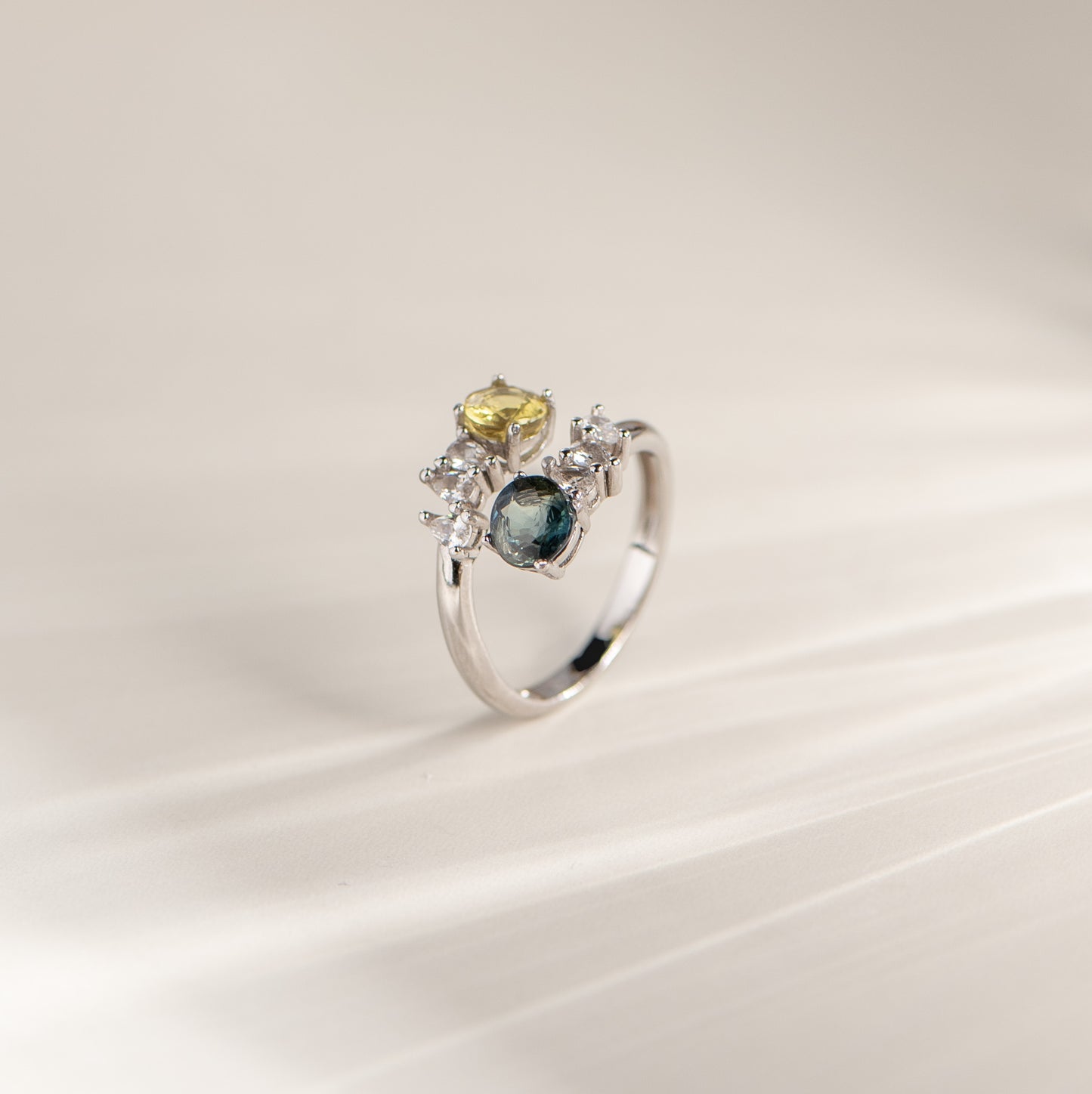 A silver ring with a cluster setting featuring a round dark blue gemstone, a smaller oval yellow gemstone, and several small clear stones, photographed on a soft cream background.