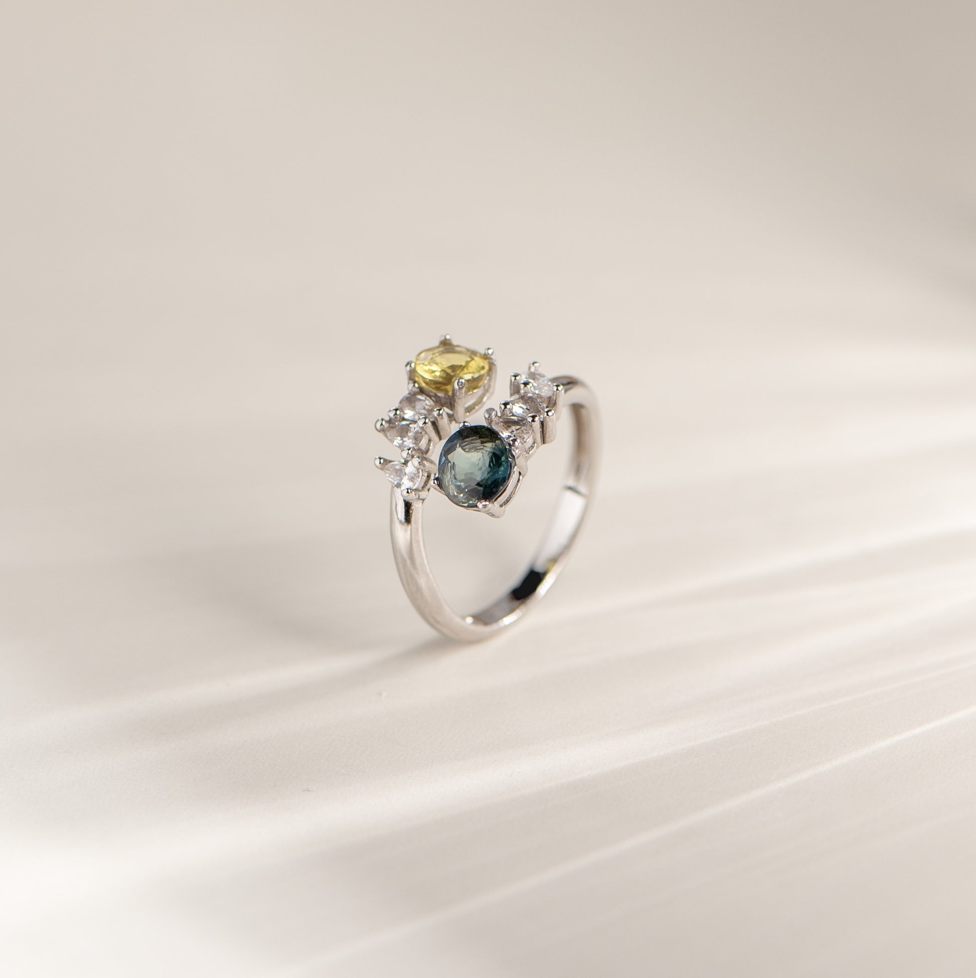 A silver ring with a cluster setting featuring a round dark blue gemstone, a smaller oval yellow gemstone, and several small clear stones, photographed on a soft cream background.