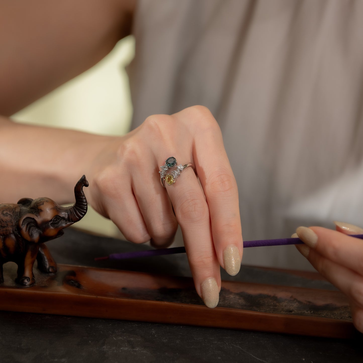 A close-up lifestyle shot of a person's hands, with one hand wearing a silver ring decorated with blue and yellow stones. The person is holding a purple pen over a wooden tray with a small elephant figurine.