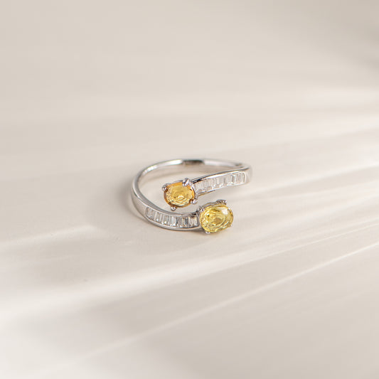 A silver-toned open ring with two oval yellow gemstones and a row of small clear stones along the band, photographed on a soft light beige surface.