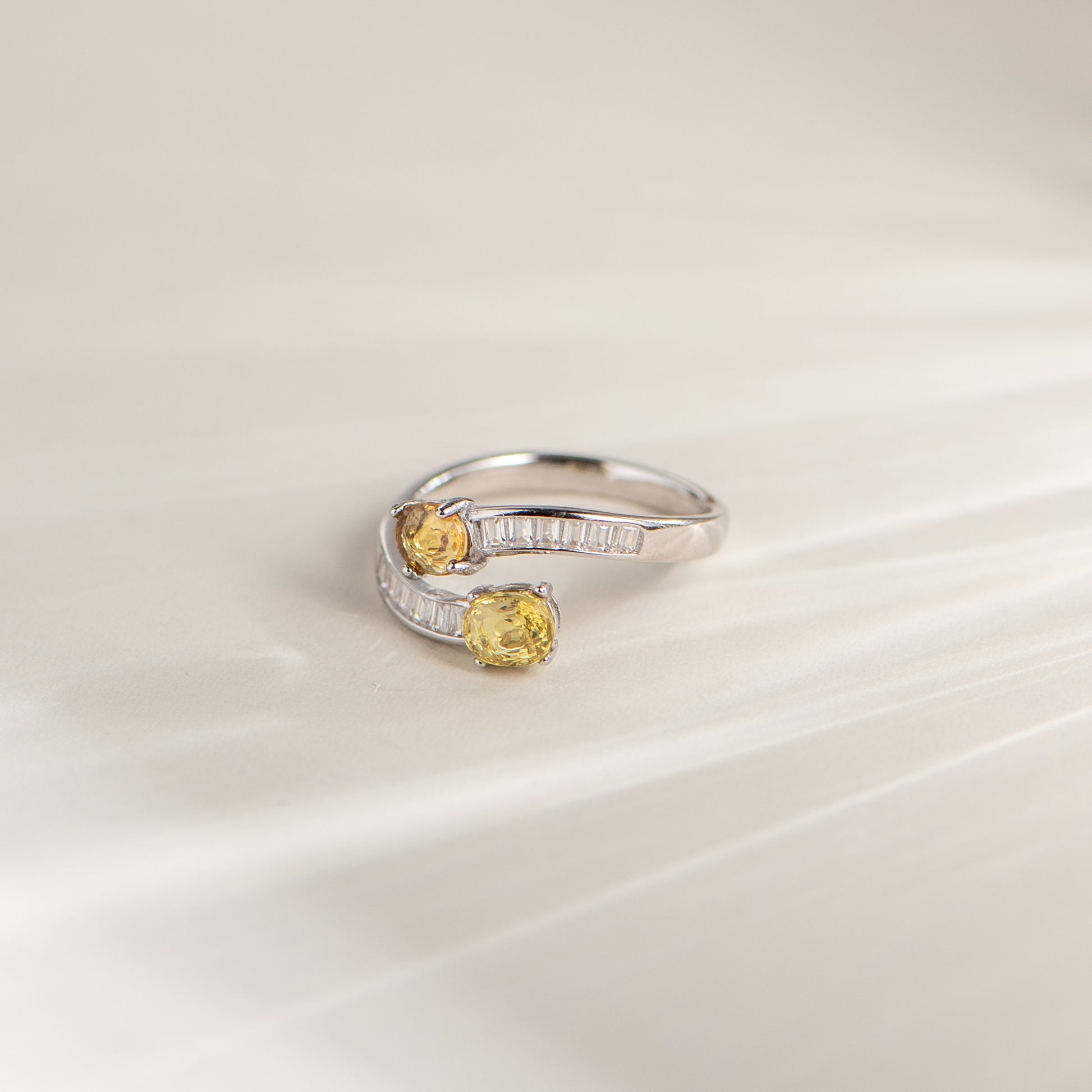 A silver wrap-style ring with two oval yellow gemstones, photographed on a soft cream surface.