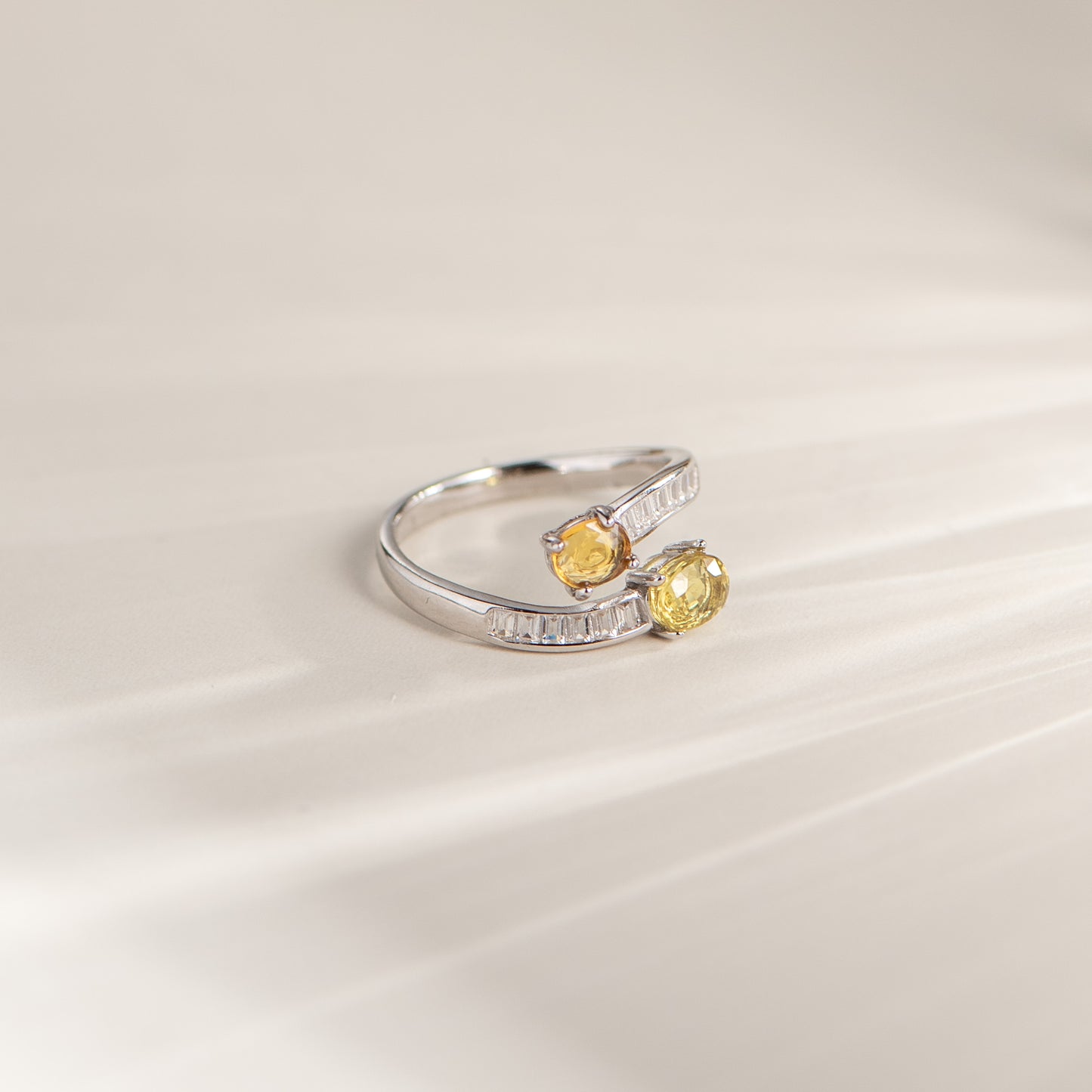 A silver-toned wrap-style ring featuring two oval yellow gemstones, photographed on a soft cream background with subtle diagonal shadows.