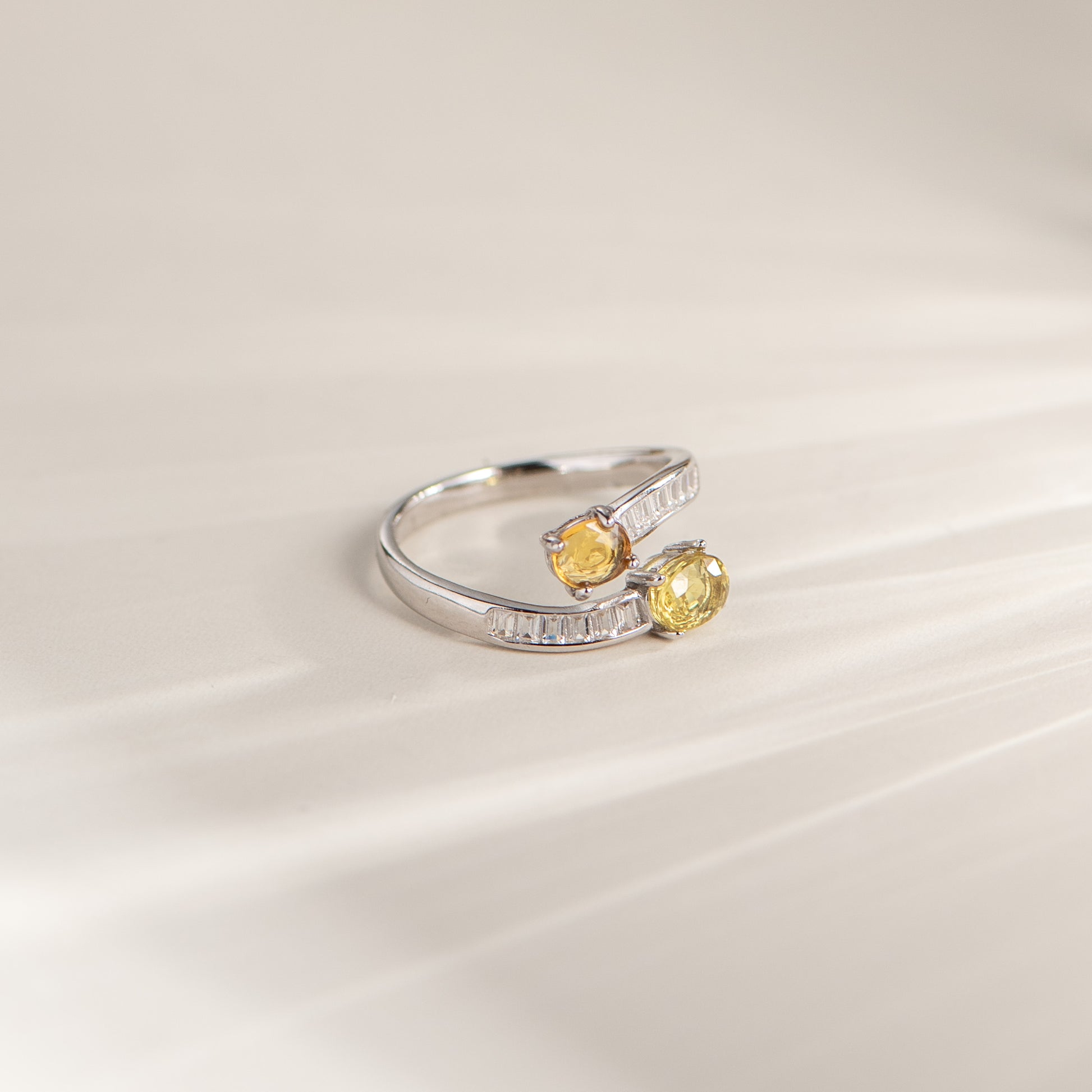 A silver-toned wrap-style ring featuring two oval yellow gemstones, photographed on a soft cream background with subtle diagonal shadows.