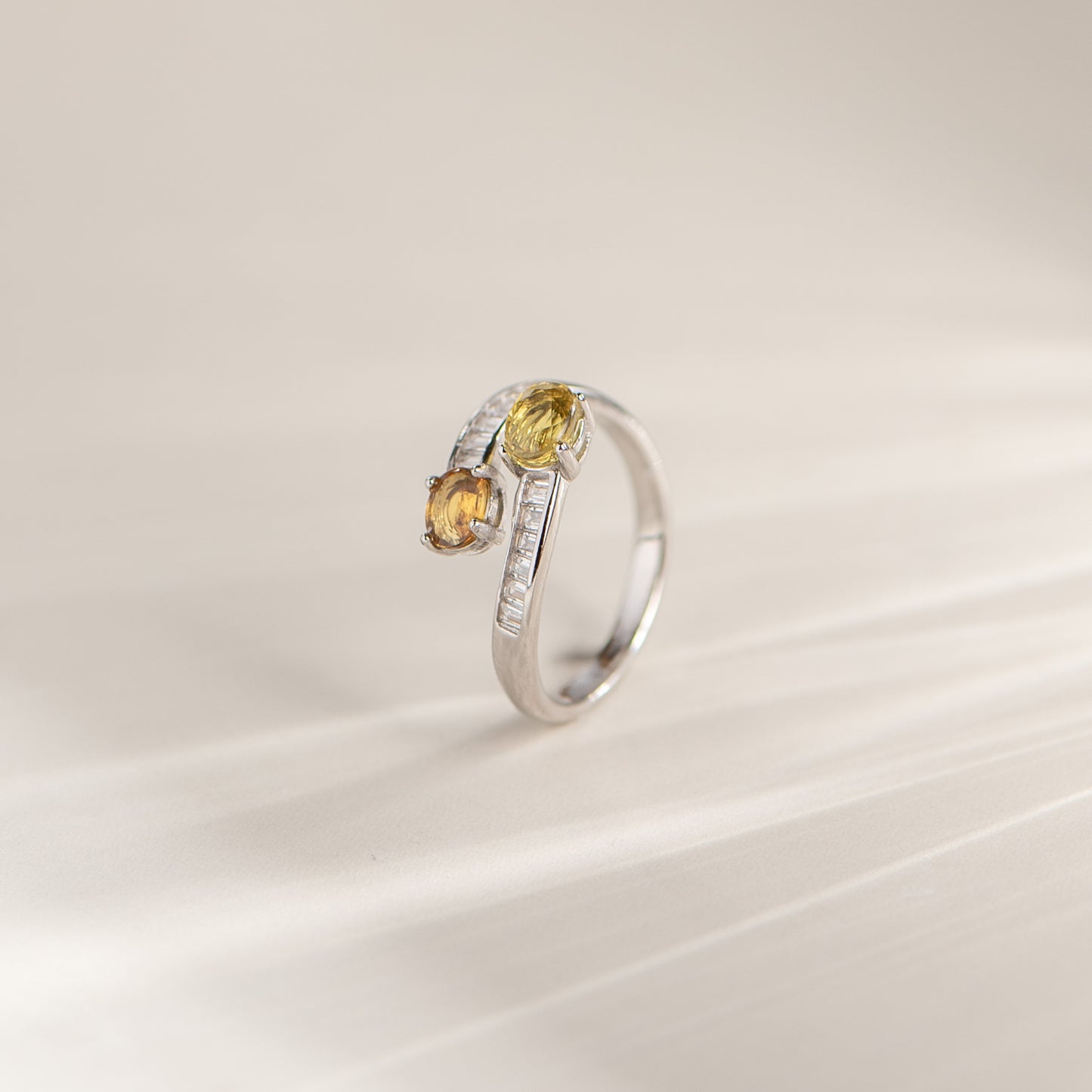 A silver ring with two yellow gemstones and a line of small clear stones, photographed on a soft cream background.
