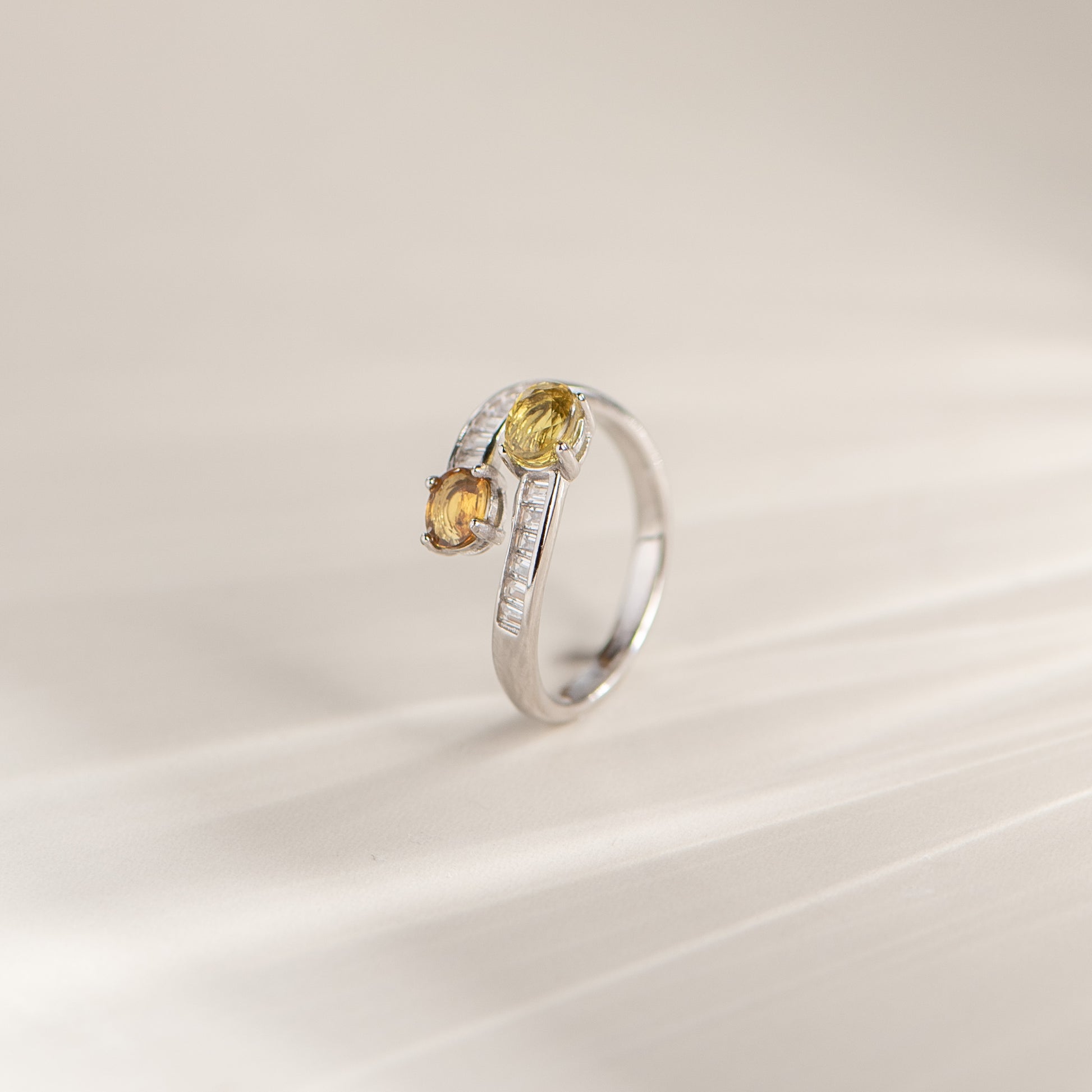 A silver ring with two yellow gemstones and a line of small clear stones, photographed on a soft cream background.