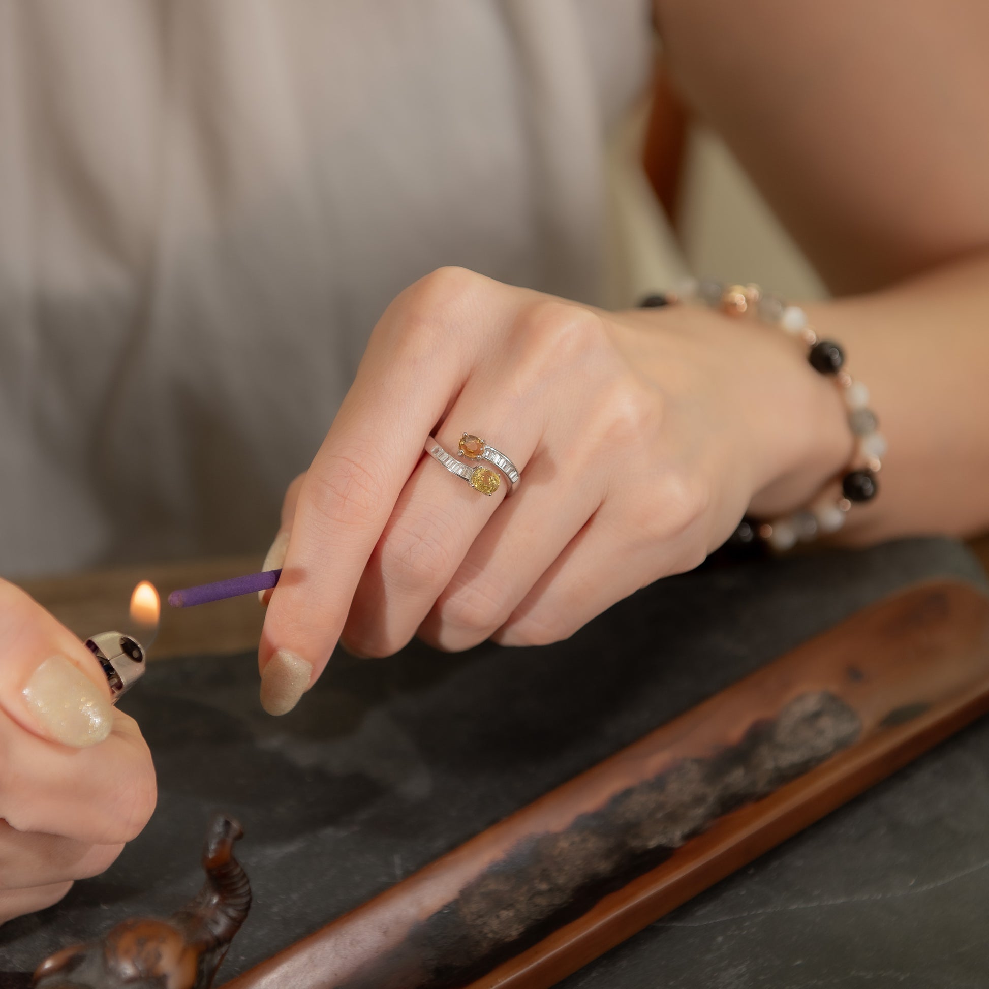 A close-up lifestyle shot of a person's hands, with one hand wearing a silver ring featuring a small yellow gemstone and clear stones, while lighting a incense stick. The person is also wearing a beaded bracelet.