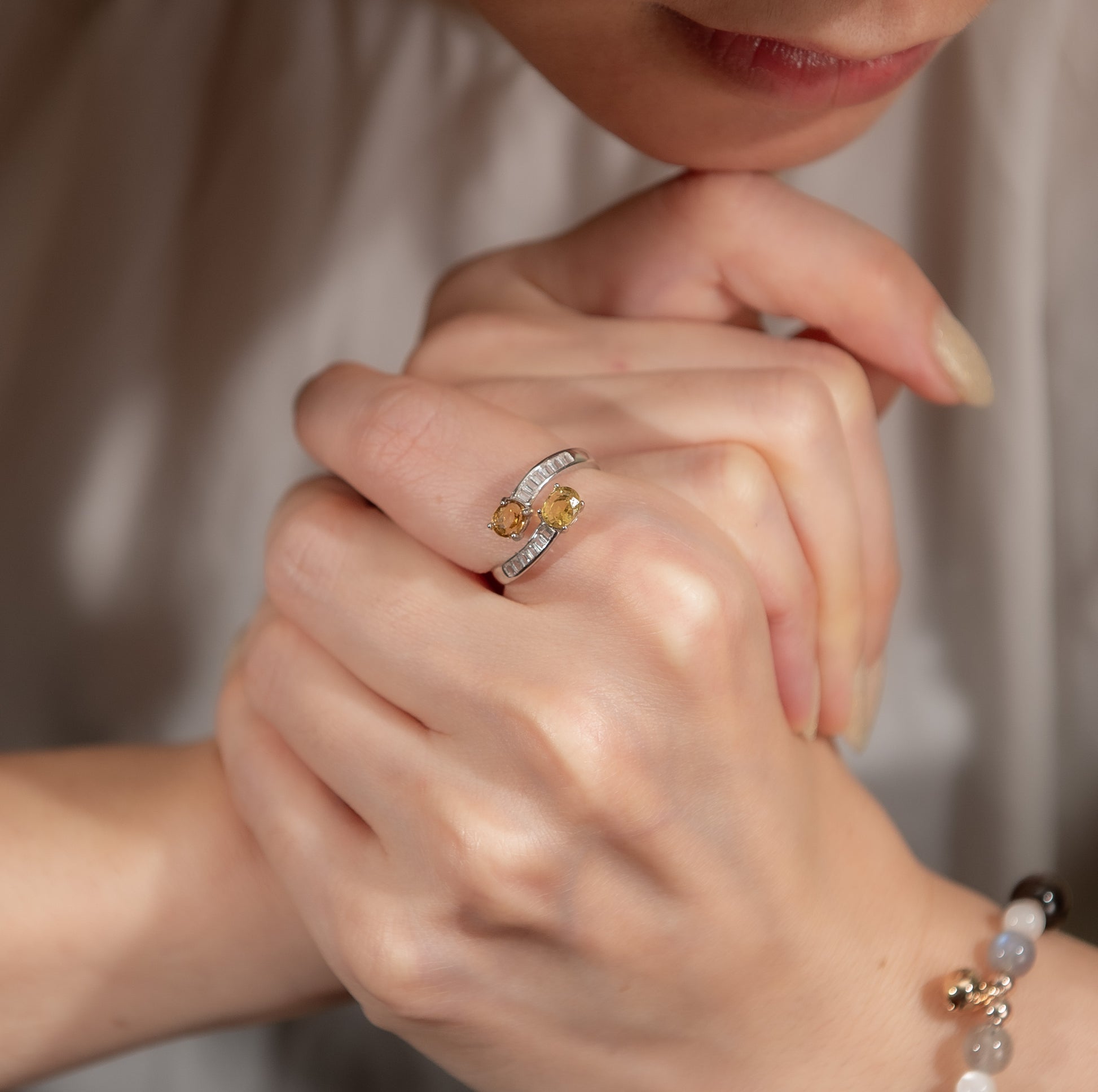A close-up lifestyle shot of a woman's hands resting together, showcasing a silver ring with two yellow gemstones on her index finger. The background is a soft, warm beige tone from her blouse.