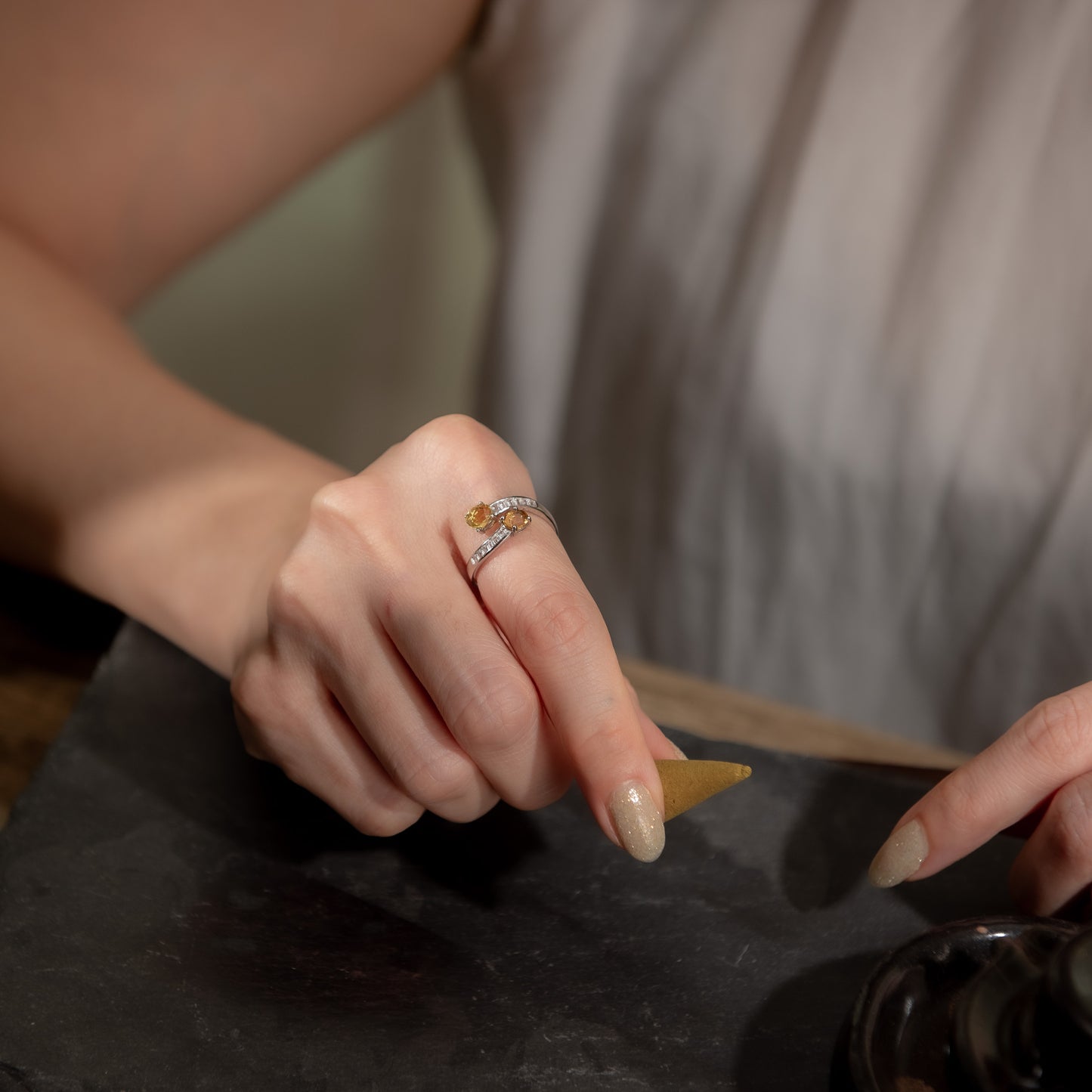A close-up lifestyle shot of a person's hand wearing a delicate silver ring with a small gold accent and clear stones, resting on a dark slate surface.