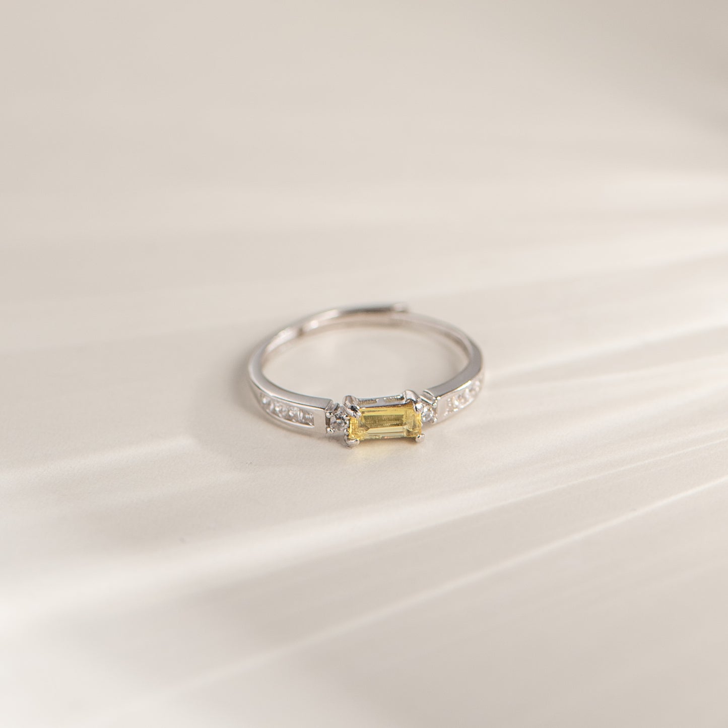 A silver ring featuring a rectangular yellow gemstone in a prong setting, accented with small clear stones along the band, photographed on a soft cream-colored surface.
