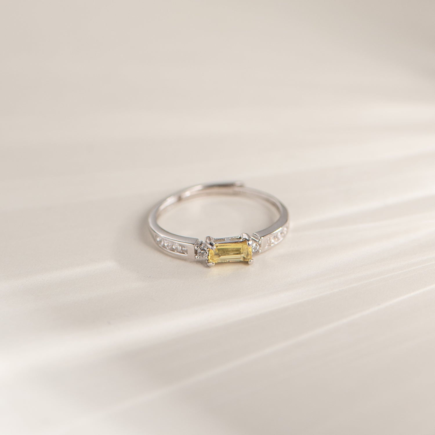 A silver ring featuring a rectangular yellow gemstone in a prong setting, accented with small clear stones along the band, photographed on a soft cream-colored surface.